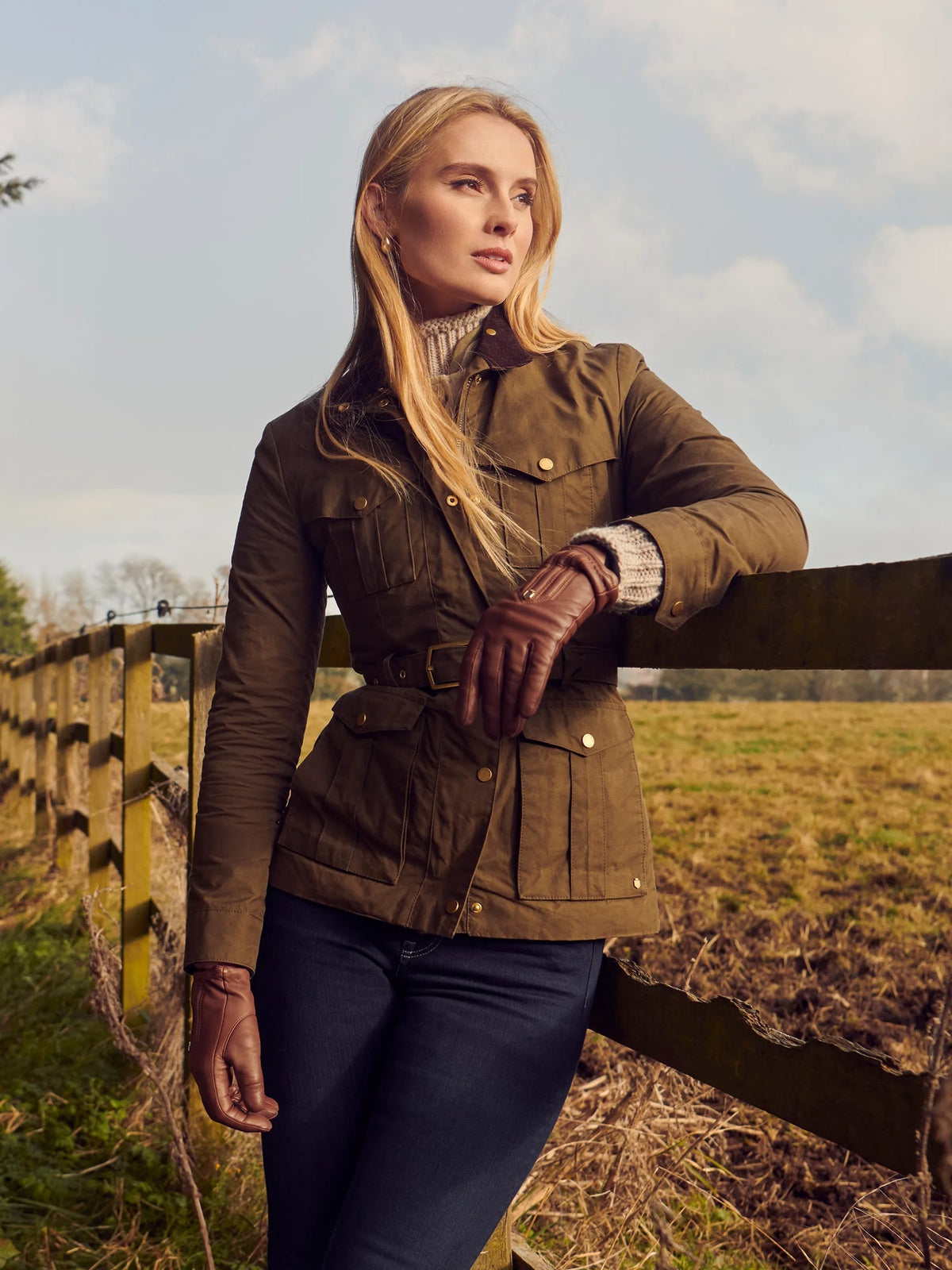 A woman stands outdoors by a wooden fence wearing a brown waxed canvas utility jacket with a matching belt, dark blue jeans, and brown leather gloves.