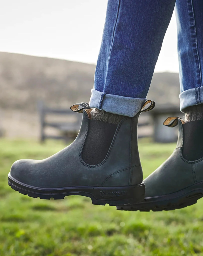 A person wearing dark gray Chelsea-style boots with elastic side panels and cuffed blue jeans, standing outdoors on grass with a blurred rural background.