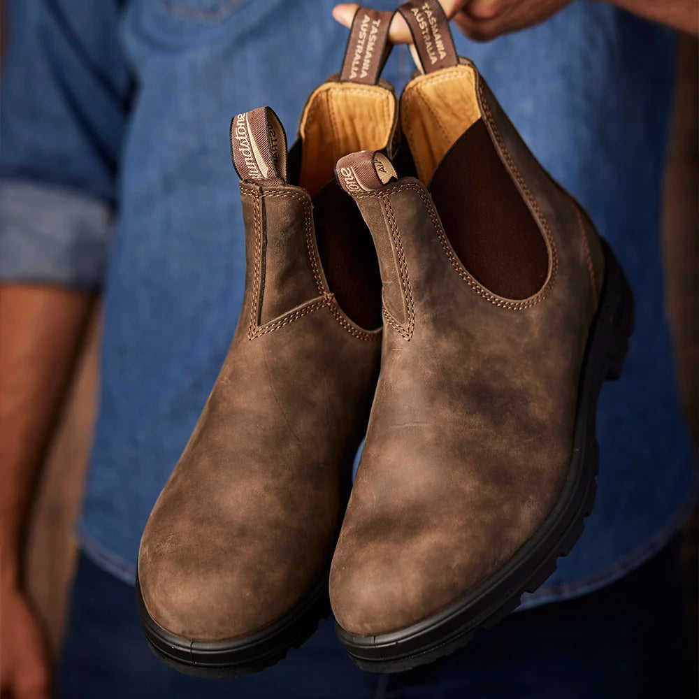 A pair of brown suede Chelsea-style boots held up by a person wearing a blue denim shirt. The boots have elastic side gore panels, pull tabs, and a rugged black sole.