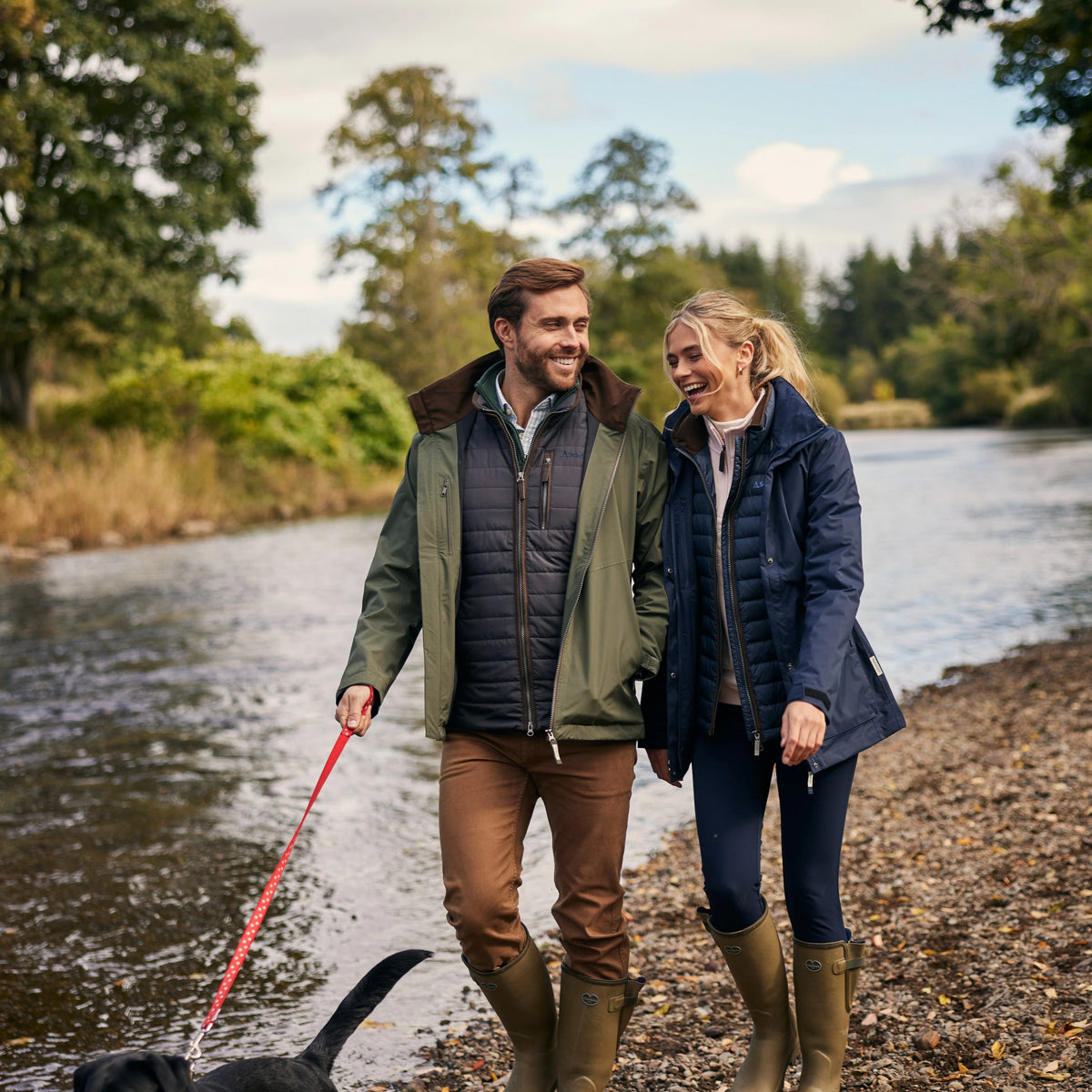 A man and a woman walking along a riverbank with a black dog on a leash. The man wears an olive green and navy puffer jacket, brown pants, and tall brown boots, while the woman wears a navy jacket, dark leggings, and tall brown boots.