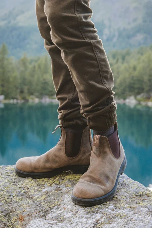 A person standing on a rock by a blue lake with forested mountains in the background, wearing brown suede Chelsea-style ankle boots and brown pants.