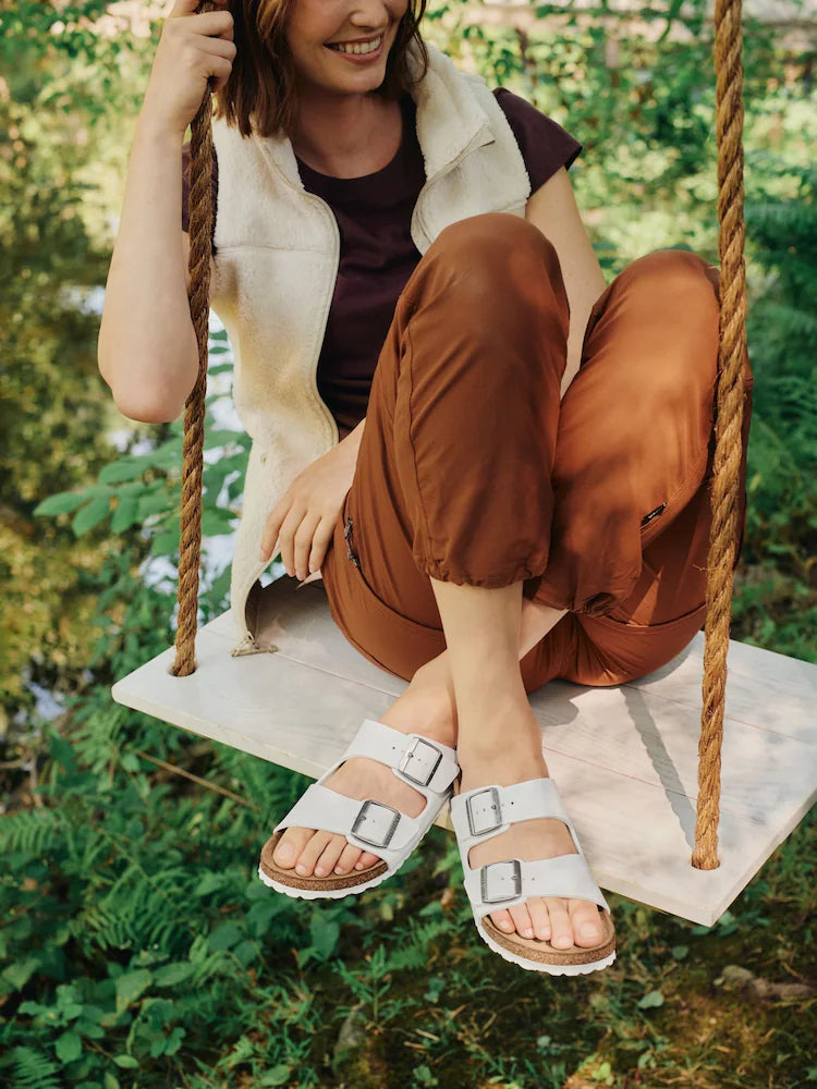 A woman sitting on a wooden swing in a lush green outdoor setting, wearing light beige double-strap sandals with square buckles, a cream sleeveless vest, and rust-brown pants.