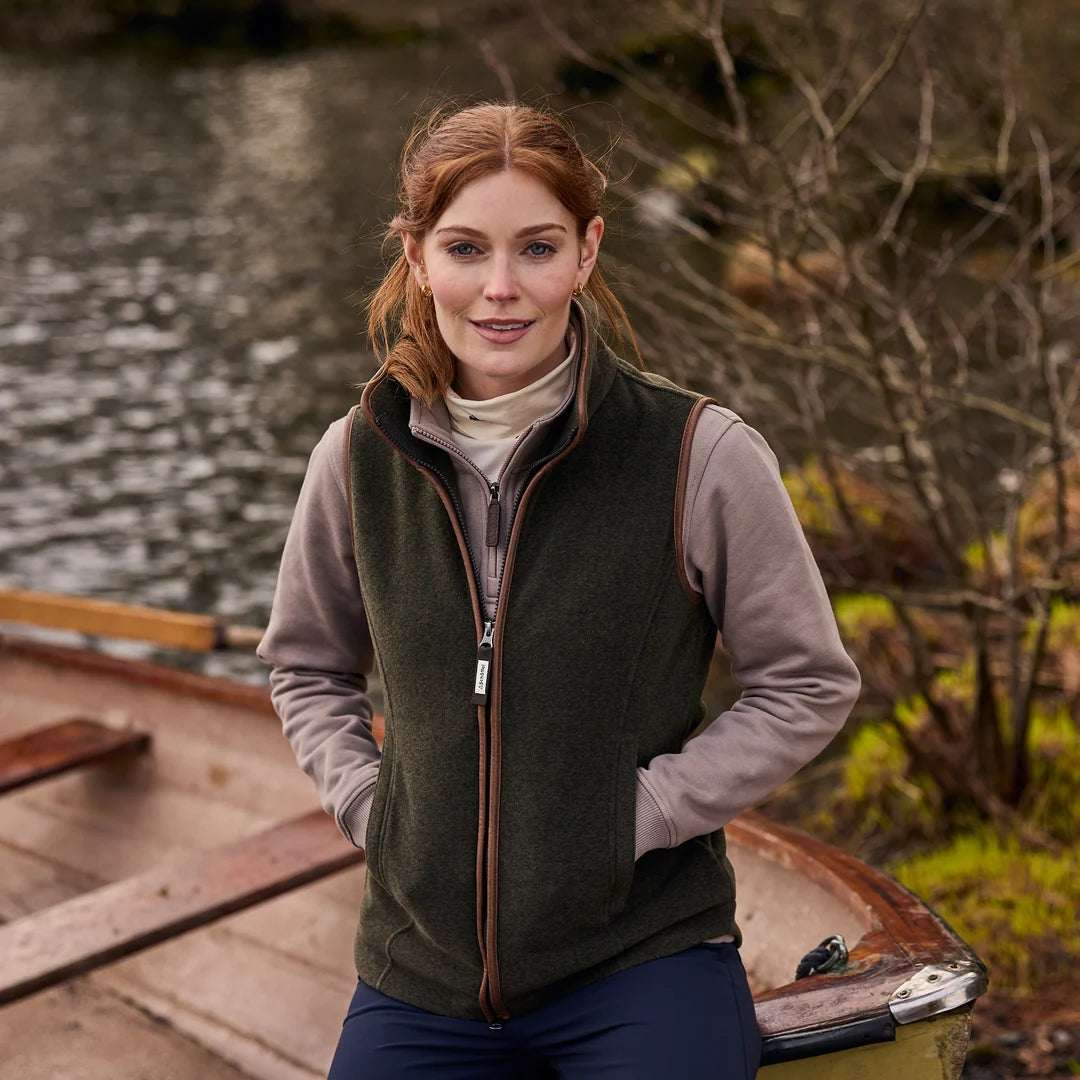 A woman wearing an olive green zip-up fleece vest over a beige turtleneck and navy pants, standing on a wooden dock next to a body of water with trees in the background.