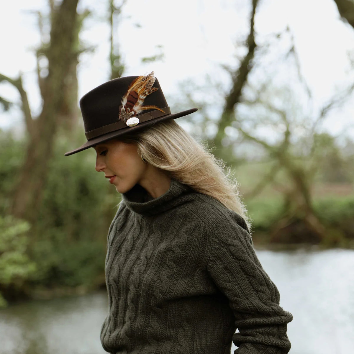 A woman in profile wearing a dark brown wide-brim hat with a feather and a dark olive green cable-knit turtleneck sweater, standing outdoors near a body of water with trees in the background.