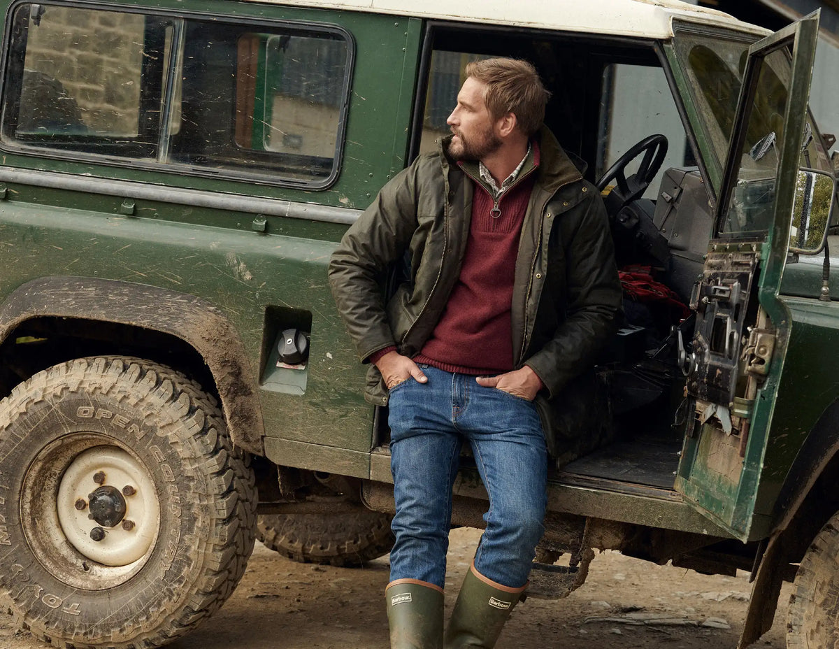A man wearing a dark olive green jacket, burgundy sweater, blue jeans, and green rubber boots stands in the doorway of a green off-road vehicle.