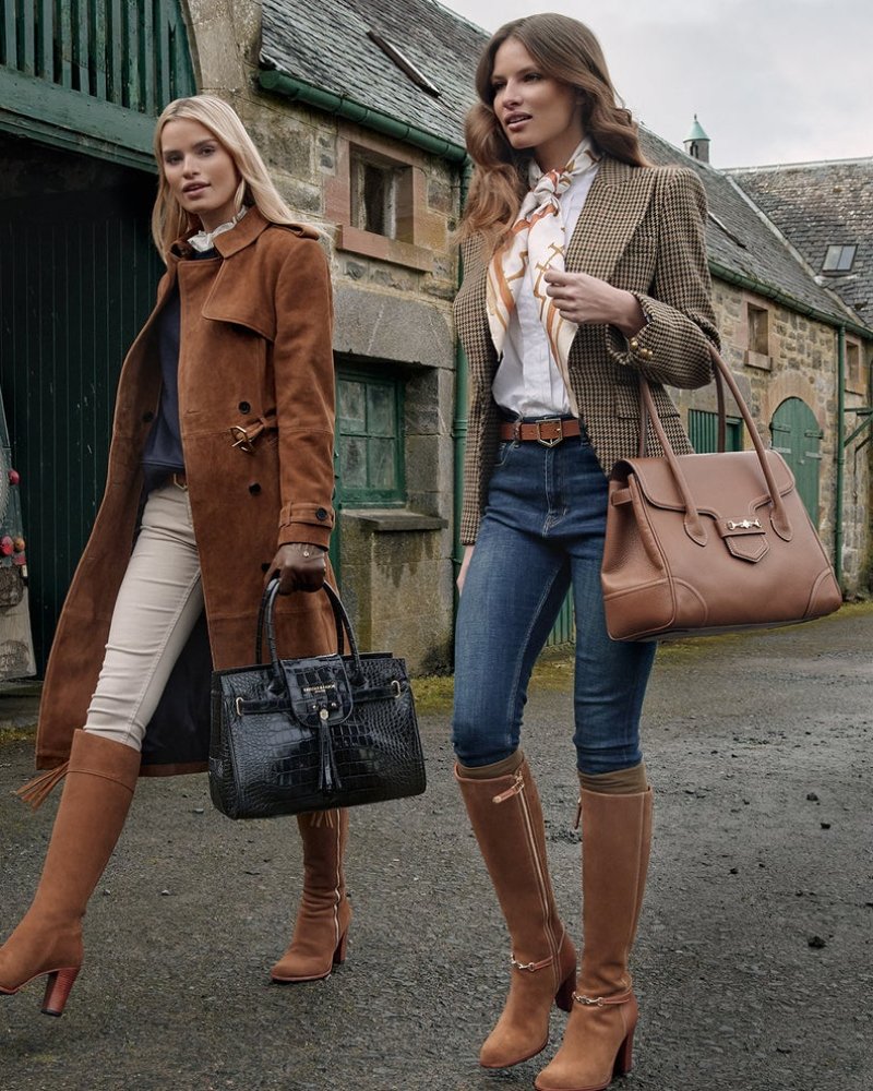 Two women standing outdoors in front of stone buildings, each carrying a large structured handbag. The woman on the left wears a brown coat, cream pants, and tan knee-high boots and carries a black crocodile-embossed handbag.